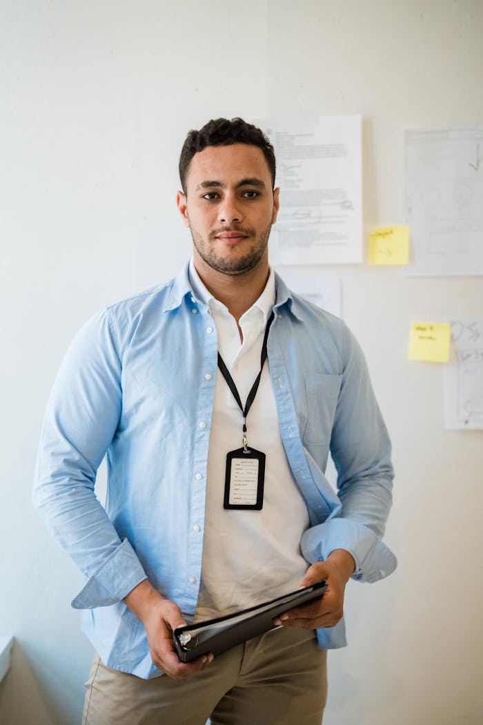 Portrait of a confident young male employee holding a notebook in an office environment with documents on the wall.